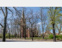 Backsteinkirche mit hohem Turm und schlankem, spitzen Kirchturm, umgeben von einem Park mit kahlen und teils austreibenden Bäumen; im Hintergrund sind vereinzelte Gebäude sichtbar.
