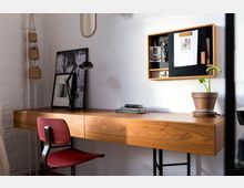 Ein minimalistischer Arbeitsplatz mit einem Holzschreibtisch, einem roten Stuhl und einer Schreibtischleuchte. An der weißen Wand hängen ein kleines Regal mit Schreibutensilien und eine Pinnwand, daneben stehen ein Blumentopf und gerahmte Bilder als Dekoration.