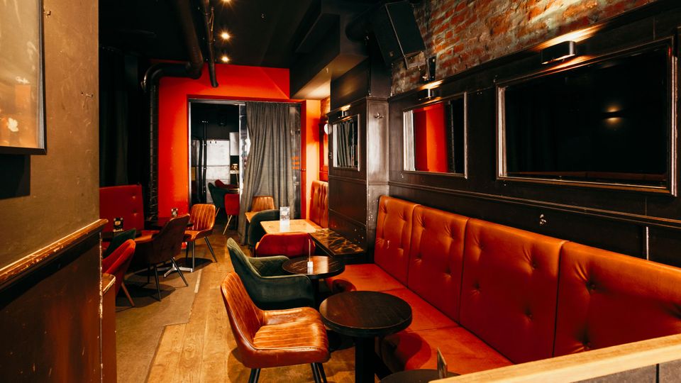 Schmaler Bar- oder Lounge-Innenraum mit roten Polsterbänken, kleinen runden Tischen und gemischten Sesseln auf Holzboden. Schwarze Wandverkleidungen mit Spiegeln, sichtbare Ziegelwand und dunkle Decke mit Spotbeleuchtung sowie ein Vorhang im hinteren Bereich prägen den Raum.