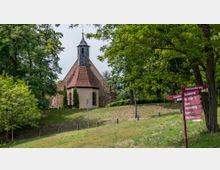 Kleine historische Kirche mit einem steinernen Turm und rotem Ziegeldach, umgeben von Bäumen und Wiese. Im Vordergrund steht ein rot beschilderter Wegweiser, der verschiedene Ziele anzeigt.