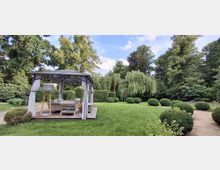 Gepflegter Gartenpark mit großer Rasenfläche, umgeben von hohen Bäumen, Hecken und kugelförmig geschnittenen Sträuchern. Links steht ein offener Pavillon mit Stoffvorhängen und Sitzmöbeln auf einer Holzplattform, rechts verläuft ein sandiger Weg.