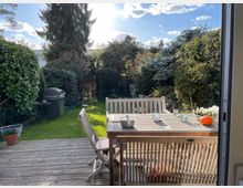 Ein kleiner Garten mit einer Holzterrasse, auf der ein Esstisch mit dazugehörigen Holzstühlen steht. Im Hintergrund sind dichte Hecken, Büsche und Bäume zu sehen, und ein Grill steht links am Rand des Rasens.