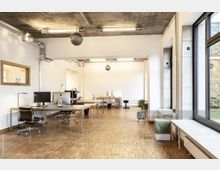 Helles, modernes Büro mit großen Fenstern, hellem Holzfußboden und schlichten weißen Wänden. Mehrere Arbeitsplätze mit Monitoren und Bürostühlen sind an schlichten Tischen verteilt, die durch minimalistische Beleuchtung und eine Sichtbetondecke ergänzt werden.