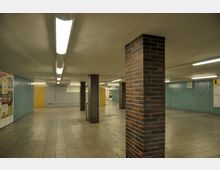 Unterirdischer Durchgangsbereich mit gefliesten Böden und Wänden, teils in blauen und gelben Tönen. Mehrere gemauerte Pfeiler aus roten Ziegeln stützen die Decke, die mit langen Leuchtstofflampen beleuchtet ist. An den Wänden befinden sich Werbeplakate und Hinweisschilder.