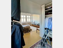 Schlafzimmer mit angrenzender Ankleide. Links sind ein offener Kleiderschrank mit hängender Kleidung und ein Anzug auf einem Bügel sowie eine Leiter sichtbar. Im Hauptbereich steht ein Bett mit einer braunen Decke, dahinter ein Sideboard vor einem großen Fenster mit mehreren kleinen Scheiben.
