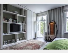 Modernes Schlafzimmer mit eingebautem weißen Regal, das Bücher, Dekor und einen Flachbildfernseher enthält. Neben einem großen Fenster mit geöffneten Doppeltüren, die auf einen Balkon führen, steht ein ovaler Spiegel; der Raum ist mit einem Teppich und Tageslicht durchfluteten Vorhängen ausgestattet.