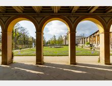 Arkaden eines historischen Gebäudes mit Blick auf einen gepflegten Garten. Im Hintergrund befinden sich Blumenbeete, ein zentraler Brunnen und ein klassizistischer Säulenpavillon, umgeben von Bäumen und weiteren Bauwerken.