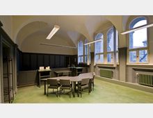 Innenraum eines historischen Gebäudes mit hohen, gewölbten Decken und großen Bogenfenstern. Der Raum ist mit grauen Holztäfelungen, grünen Heizkörpern, einem grünen Fußboden und mehreren Tischen und Stühlen ausgestattet.