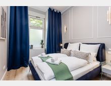 Modernes Schlafzimmer mit Doppelbett in weißer Bettwäsche, blauem Polsterbettgestell und grünem Bettläufer; an den Wänden sind Zierleisten und schlichte Wandlampen. Ein Fenster mit dunkelblauen Vorhängen, Heizkörper darunter sowie ein kleiner Beistelltisch aus Glas ergänzen den Raum.