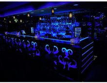 Eine moderne Bar mit blauer Neonbeleuchtung, verziert mit dekorativen Mustern an der Front des Tresens. Im Hintergrund befinden sich eine gut bestückte Getränkeregale, ein Fernseher und Hängelampen mit warmem Licht.