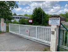 Geschlossene weiße Schranke am Eingang eines Betriebsgeländes mit umliegenden Bäumen und einem flachen Industriegebäude im Hintergrund. Schilder weisen auf Zutrittsbeschränkungen und eine Paketdienstanweisung hin.