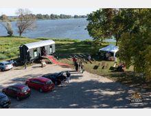 Ein Uferbereich mit Blick auf einen Fluss, im Vordergrund ein gepflasterter Parkplatz mit Autos. Rechts steht ein weißes Pavillonzelt, links ein umgebauter, schwarzer Eisenbahnwagen mit rotem Teppich am Eingang, umgeben von Menschen und Bänken auf einer Grünfläche.