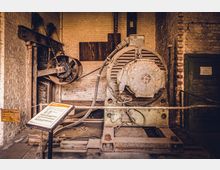 Industriehistorischer Raum mit einer großen alten Maschine aus Metall, bestehend aus einem Motor und einem Riementrieb, vor einer Backsteinwand. Im Vordergrund steht ein Schild mit Informationen über die Anlage.