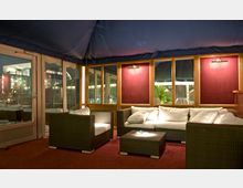 Gemütlicher Lounge-Bereich mit Rattanmöbeln und weißen Polstern, umgeben von Holzpaneelen mit großen Fenstern und dunkelroten Wandflächen, beleuchtet von warmem Licht. Im Hintergrund ist durch die Fenster ein beleuchtetes Gebäude und nächtliche Vegetation sichtbar.