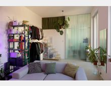 Helles Wohnzimmer mit Sofa im Vordergrund, offenem Kleiderregal und Aufbewahrungsboxen an der linken Wand sowie kleiner Treppe zu einer erhöhten Nische. Rechts trennt eine hohe, halbtransparente Glaswand den Raum; große Fenster und mehrere Zimmerpflanzen ergänzen die moderne Einrichtung.