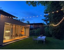 Ein moderner Bungalow mit großen Glasfronten und heller Innenbeleuchtung, umgeben von einem Garten mit Rasen. Im Vordergrund steht ein gedeckter Tisch mit Stühlen, darüber hängen Lichterketten, die in der Dämmerung leuchten.