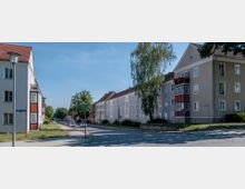 Straßenszene mit mehrstöckigen Wohnhäusern in heller Putzfarbe und roten Giebeldächern, umgeben von Bäumen und Wiesenflächen. Die Straße besteht aus Pflastersteinen, mit parkenden Autos und einer Straßenlaterne im Vordergrund.