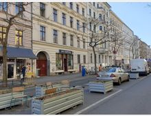 Straßenszene mit historischen Wohnhäusern in heller Putzfassade, Geschäften im Erdgeschoss wie einem Café (Schwesterherz) und einer Boutique (ete clothing). Autos parken entlang der Straße, die mit Bäumen und rechteckigen Pflanzkübeln gesäumt ist.