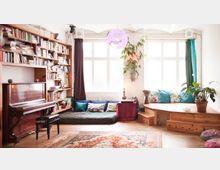 Helles Wohnzimmer mit einer großen Bücherwand, einem Klavier und Sitzbereich. Im Vordergrund ein orientalischer Teppich, im hinteren Bereich Matratzen mit Kissen und eine hölzerne Stufensitzfläche, dekoriert mit Pflanzen und einer Hängelampe.