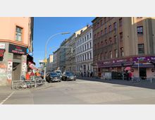 Straßenecke in einer städtischen Umgebung mit mehrstöckigen Wohn- und Geschäftsgebäuden. Links ist ein Sushi-Restaurant mit einem schwarzen Schild zu sehen, rechts ein Blumenladen mit einer lilafarbenen Markise; Autos und Fahrräder stehen am Straßenrand.