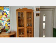 Innenbereich eines Flurs mit einem hohen Holz-Vitrinenschrank mit Glastüren, daneben eine helle Tür mit drei schmalen Milchglasfenstern. Links ist ein großes, buntes Buntglasfenster zu sehen sowie ein kleiner Beistelltisch mit Gegenständen.