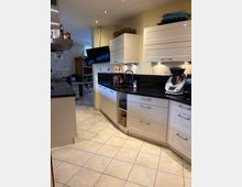 Moderne Küche mit weißen Schränken, schwarzen Arbeitsplatten und Einbauleuchten unter den Hängeschränken. Im Vordergrund sind Küchenutensilien und ein Küchenmaschine sichtbar, während im Hintergrund ein Flachbildfernseher und ein Bücherregal stehen.