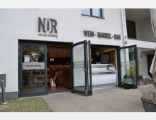 Eingangsbereich eines Weinhandels mit Bar („NUR – not only riesling“) in einem modernen, hell verputzten Gebäude, mit großen Glastüren und aufgesetzter Beschriftung „WEIN · HANDEL · BAR“. Durch die offenen Türen sind Theke und Kühlvitrine im Innenraum sichtbar; davor liegt eine gepflasterte Terrasse mit Sitzmöbeln.
