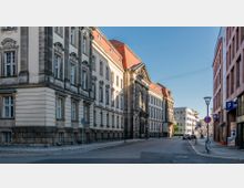 Straßenansicht mit einem historischen Gebäude auf der linken Seite, das weiße Fassaden, verzierte Fensterrahmen und ein rotes Dach zeigt. Rechts befinden sich moderne Gebäude und Geschäfte mit Parkschildern, die eine Mischung aus Alt- und Neubauten in einer städtischen Umgebung zeigen.