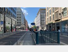 Straßenansicht in einer Stadt mit modernen und klassischen Gebäuden, einem U-Bahn-Eingang mit blauer Beschilderung "U Stadtmitte" im Vordergrund und erkennbaren Einzelhandelsgeschäften auf beiden Straßenseiten. Fußgänger und Fahrradbügel befinden sich entlang des gepflasterten Gehwegs.