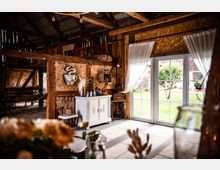 Innenraum eines rustikalen Gebäudes mit Holzbalken, einer weißen Anrichte, dekorativem Zubehör und einer großen Glastür mit Blick auf einen grünen Außenbereich. Licht strömt durch transparente Vorhänge und erhellt den Raum.