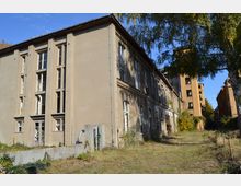 Abgebildet ist ein verlassenes, mehrstöckiges Gebäude mit verputzter Fassade und großen rechteckigen Fenstern, die teilweise beschädigt sind. Im Hintergrund befinden sich weitere alte, teils überwucherte Gebäude, umgeben von einer Wiese mit vereinzelten Bäumen.