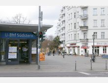 Eingang des U-Bahnhofs Südstern in Berlin, mit einem kleinen Kiosk und Hinweisschildern an der Tür, daneben eine Straße mit Ampel. Im Hintergrund ein Platz mit Cafés, Sitzbereichen und Wohnhäusern im Altbaustil.