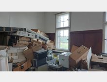 Ein Raum mit hohen Fenstern und Holzvertäfelung an den Wänden, der mit gestapelten Kartons, Computergehäusen und diversen Elektroartikeln unordentlich gefüllt ist. Durch das Fenster ist ein Blick auf eine grüne Außenfläche sichtbar.