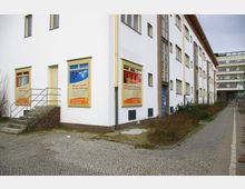 Mehrstöckiges Wohngebäude im Stil der Nachkriegsmoderne mit weißer Fassade und gelben Fensterrahmen. Vor dem Gebäude befindet sich ein gepflasterter Gehweg, Werbeplakate an den Fenstern werben für Wohnungsangebote.