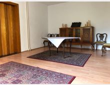 Ein schlicht eingerichteter Raum mit hellem Holzboden, einem rechteckigen Tisch mit Tischdecke, umgeben von klassischen, gepolsterten Stühlen. An der weißen Wand steht eine verzierte Holzvitrine, dahinter befinden sich dekorative Holzobjekte; der Boden ist mit orientalen Teppichen bedeckt.