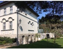 Historisches Gebäude mit hellem Putz und dekorativen Elementen, darunter Rundbogenfenster und ein steinernes Fundament. Im Vordergrund ein gepflegter Garten mit Bäumen, Sträuchern und einer blühenden Wiese.