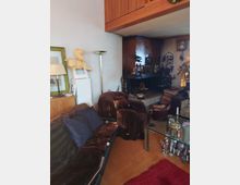 Wohnzimmer mit Holzboden, Sofas mit braunen Überwürfen, einem Kamin in einer Ecke aus dunklem Holz und dekorativen Figuren. Ein Couchtisch aus Glas steht auf einem roten Teppich, und Wände sind mit Bildern und verschiedenen Dekorationen geschmückt.