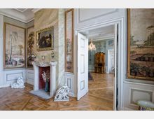 Eleganter Innenraum eines historischen Zimmers mit dekorativem Kamin, verziertem Rahmenbild und antikem Mobiliar. Wandgemälde, Marmorelemente und hölzerner Parkettboden prägen das Ambiente; eine offene Tür führt zu einem weiteren Raum mit Kronleuchter und Schrank.