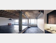 Großer, loftartiger Raum mit Betonboden und freiliegender Holzdecke, einer gefliesten Säule in der Mitte, großen Fenstern, die Tageslicht einlassen, einer kleinen erhöhten Plattform im Hintergrund und einer Küchenzeile mit Waschbecken und Herd auf der rechten Seite. Ein Sofa und ein Studiolicht stehen in der linken Ecke.