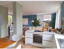 Ein modernes, offenes Wohnzimmer und eine Küche mit heller Einrichtung. Die Küche hat eine weiße Kochinsel mit integriertem Herd und Hängelampen, während das Wohnzimmer mit einer blauen Akzentwand, Pflanzen und gemütlichen Möbeln ausgestattet ist.