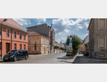 Straßenszene in einer historischen Altstadt mit gepflasterter Fahrbahn, flankiert von farbigen Stadtvillen und historischen Gebäuden mit roten Ziegeldächern. Am Straßenrand sind parkende Autos, kleine Bäume, Laternen und Schilder sichtbar, während ein sonniger Himmel die Szenerie erhellt.