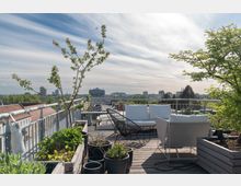 Dachterrasse mit Holzboden, bepflanzten Blumenkästen und moderner Sitzgruppe, umgeben von Geländern und mit Blick auf eine Stadtlandschaft mit niedrigen Gebäuden und Hochhäusern im Hintergrund. Ein Baum und weitere Pflanzen sorgen für Begrünung, während am Himmel leichte Wolken zu sehen sind.