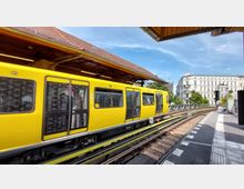 Gelbe U-Bahn an einem erhöhten Bahnhof mit überdachtem Bahnsteig und sichtbaren Gleisen, im Hintergrund ein mehrstöckiges historisches Gebäude und blauer Himmel. Ein Bereich für Rollstuhlfahrer ist an der Wagontür gekennzeichnet.