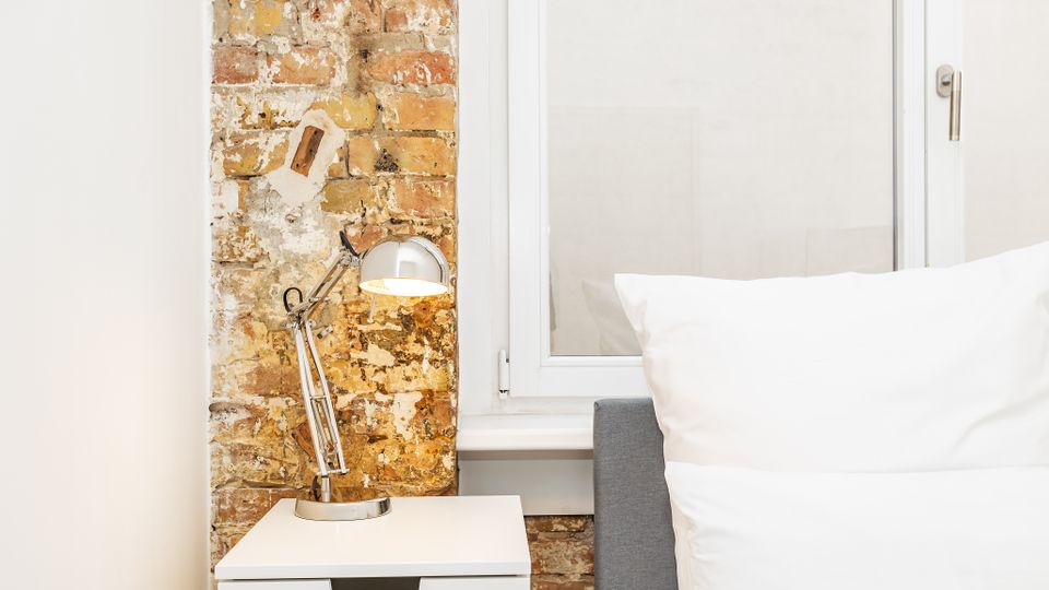 Schlafzimmer-Ecke mit einem Bett mit weißen Kissen neben einem weißen Nachttisch und einer verstellbaren Metall-Schreibtischlampe. Dahinter sind eine freigelegte Ziegelwand und ein großes Fenster mit weißem Rahmen zu sehen.
