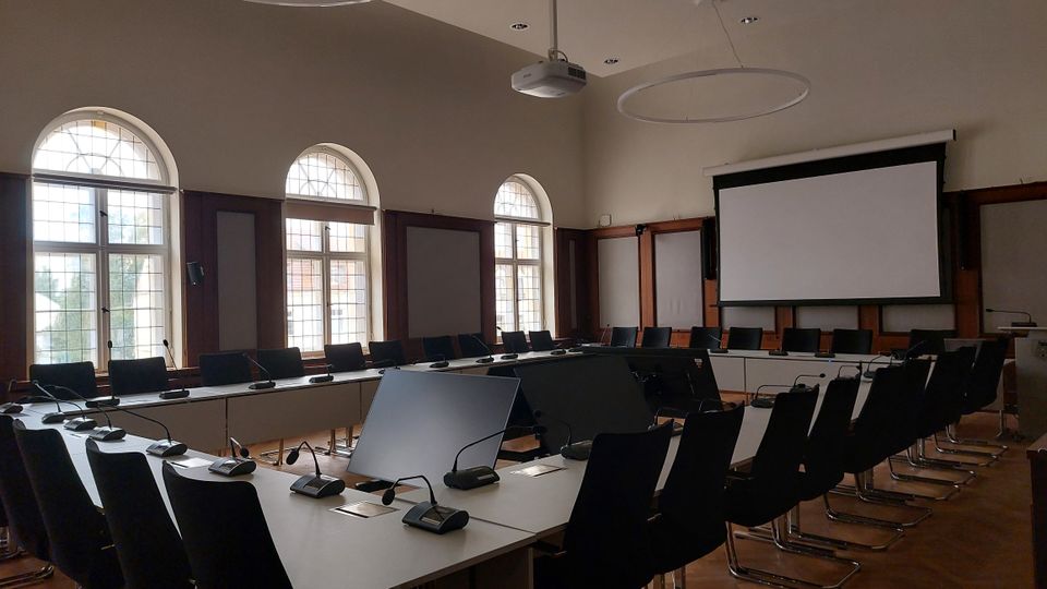 Konferenzraum mit langen, rechteckigen Tischen in U-Form, umgeben von schwarzen Stühlen. Der Raum hat hohe Fenster mit Rundbögen, eine Leinwand an der Stirnseite und moderne Mikrofone auf den Tischen.