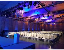 Veranstaltungsraum mit industriellem Charme, gekennzeichnet durch unverputzte Ziegelwände und hohe Decken mit Scheinwerfern und dekorativen Beleuchtungselementen. Reihen aus weißen Stühlen und ein blauer Bühnenbereich sind vorbereitet, unterstützt von einer stimmungsvollen blauen Beleuchtung.