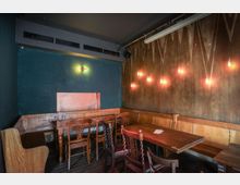 Ein kleiner Innenraum mit Holzbänken und Holztischen, umgeben von dunkelgrünen und braunen Wänden. An der Wand hängen mehrere glühende, freihängende Glühbirnen, die in warmem Licht leuchten.