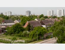 Wohnsiedlung mit einer Mischung aus Einfamilienhäusern mit Giebeldächern und viel grüner Vegetation im Vordergrund; im Hintergrund stehen hohe, moderne Wohnblöcke und ein Funkturm ist in der Ferne erkennbar.