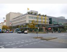 Straßenszene mit einem großen, mehrstöckigen Kaufhaus der Marke „Karstadt“ im Zentrum des Bildes, umgeben von einer breiten Straße mit wenigen Autos. Das Gebäude hat eine helle Steinfassade, viele Fenster und eine bepflanzte Dachterrasse, daneben steht ein kleiner Pavillon mit dunklem Dach.