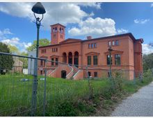 Rötliche Villa im klassizistischen Stil mit Rundbögen, einem quadratischen Turm und großen Fenstern, umgeben von einem grünen Rasen und Bauzäunen. Im Vordergrund steht eine ältere Straßenlaterne, daneben ein gepflasterter Gehweg unter blauem Himmel mit weißen Wolken.
